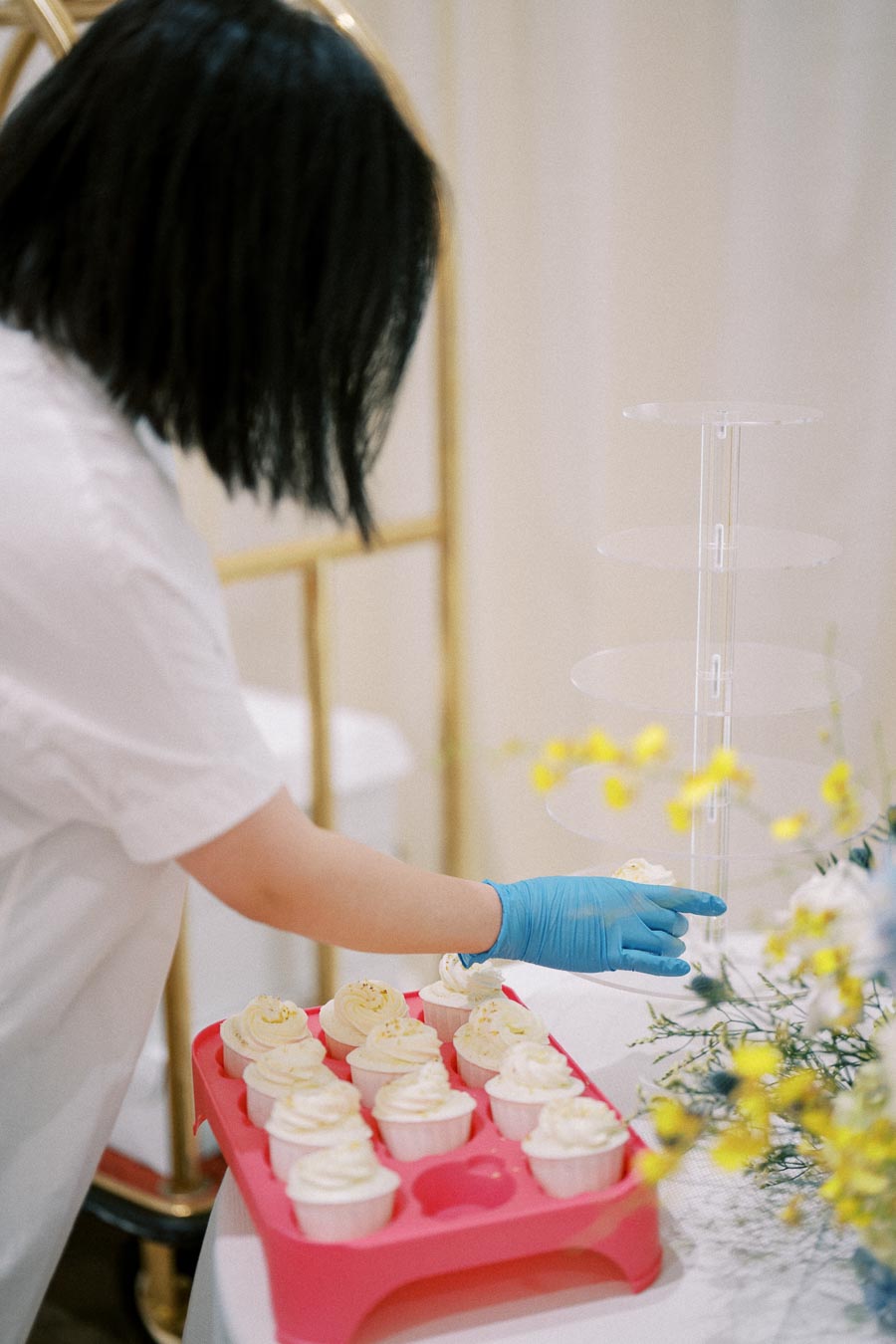 A person arranging a tray of vanilla cupcakes with frosting on a table, wearing blue gloves, next to a clear cupcake stand and yellow flowers in the foreground.