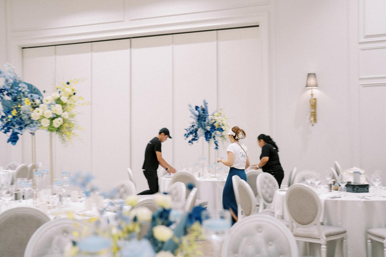 Elegant banquet hall setup with staff preparing tables, featuring white tablecloths and blue floral centerpieces.