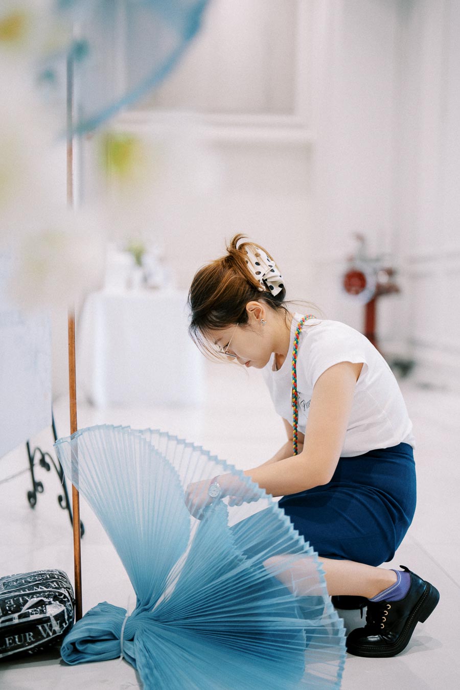Young woman crouching indoors, focusing on folding a large blue fabric, with a vintage bag nearby.
