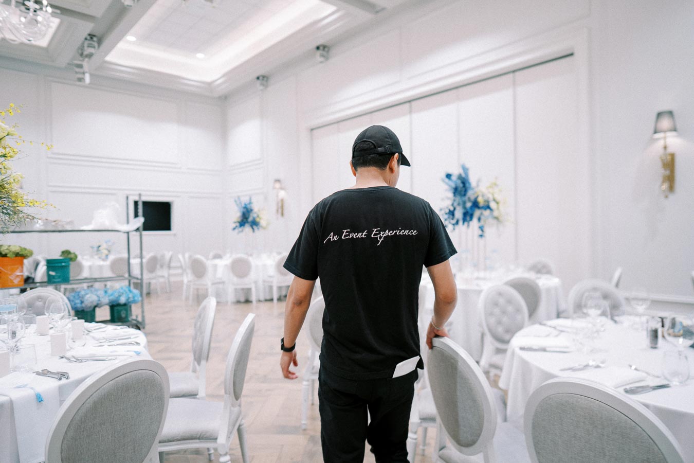 A person in a black shirt with An Event Experience on the back setting up a luxurious event space with elegantly decorated white tables and chairs.