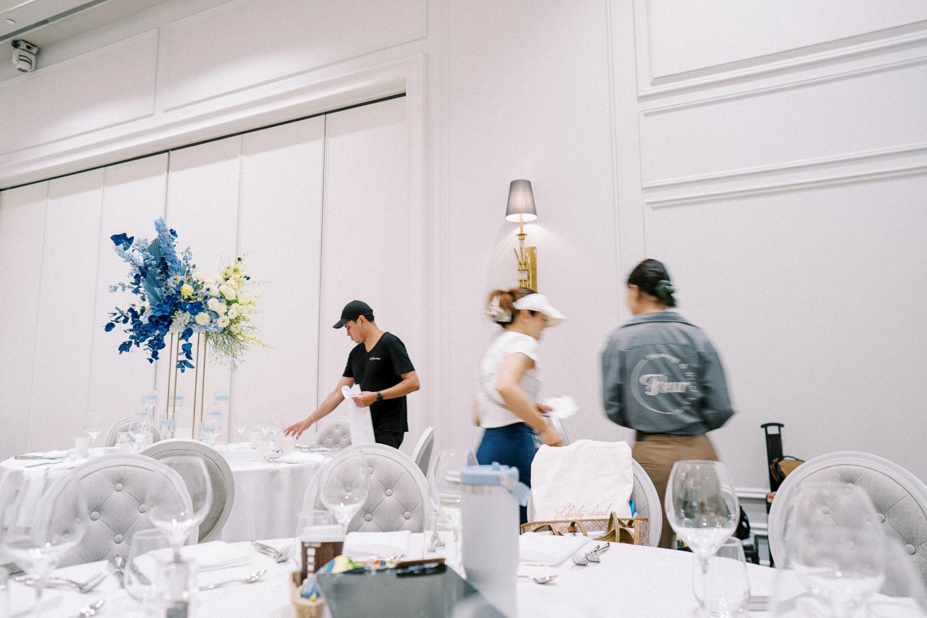 Elegant banquet setup with staff preparing tables, featuring white tablecloths, ornate flower arrangements in blue and yellow, and plush chairs in a sophisticated dining venue.