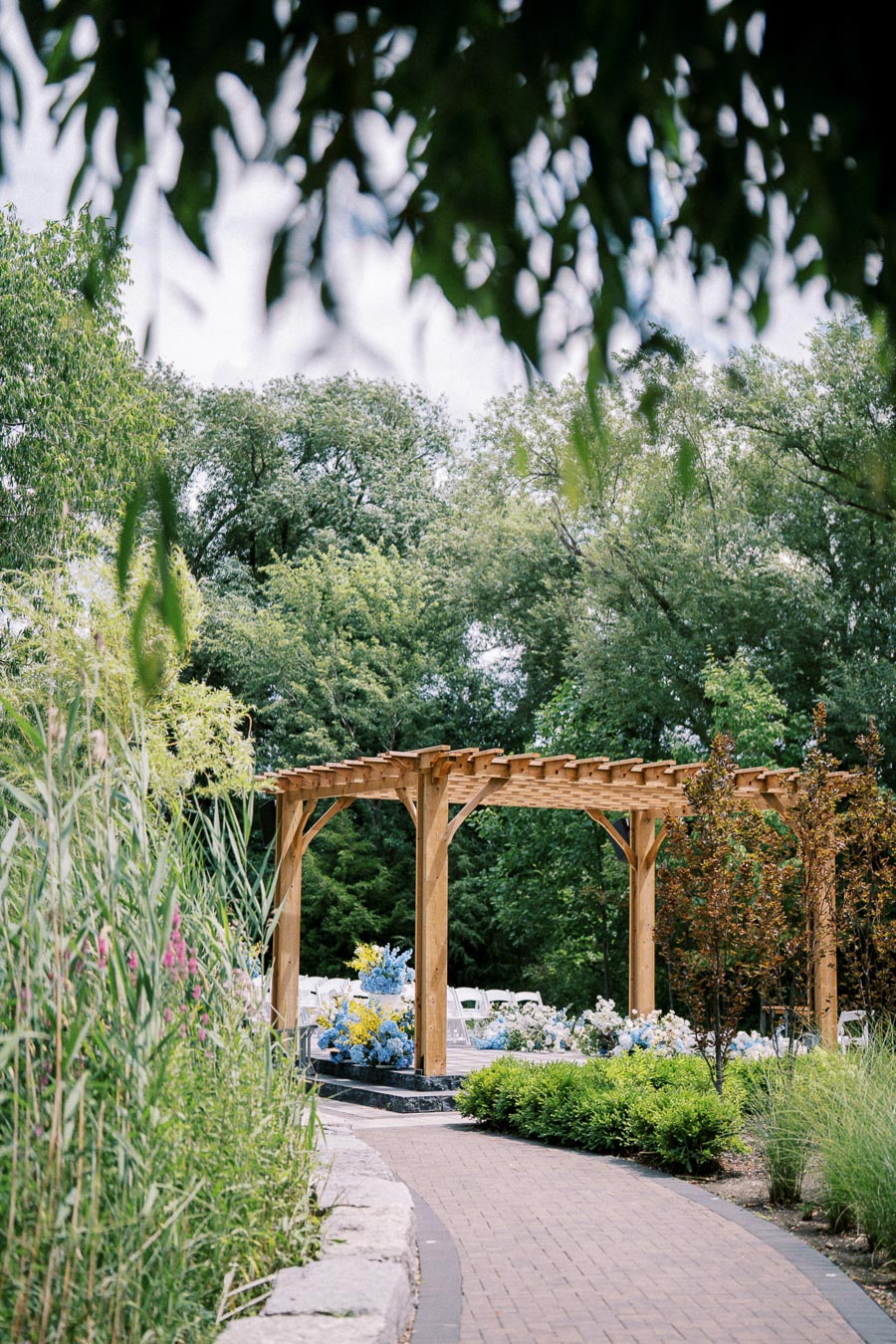 Wooden pergola in a lush garden setting with a brick pathway, surrounded by green trees and colorful flowers.