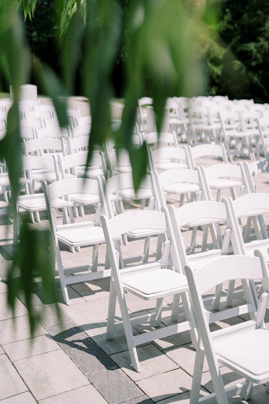 Rows of white folding chairs set up outdoors for a wedding ceremony, partially obscured by green leaves in the foreground.