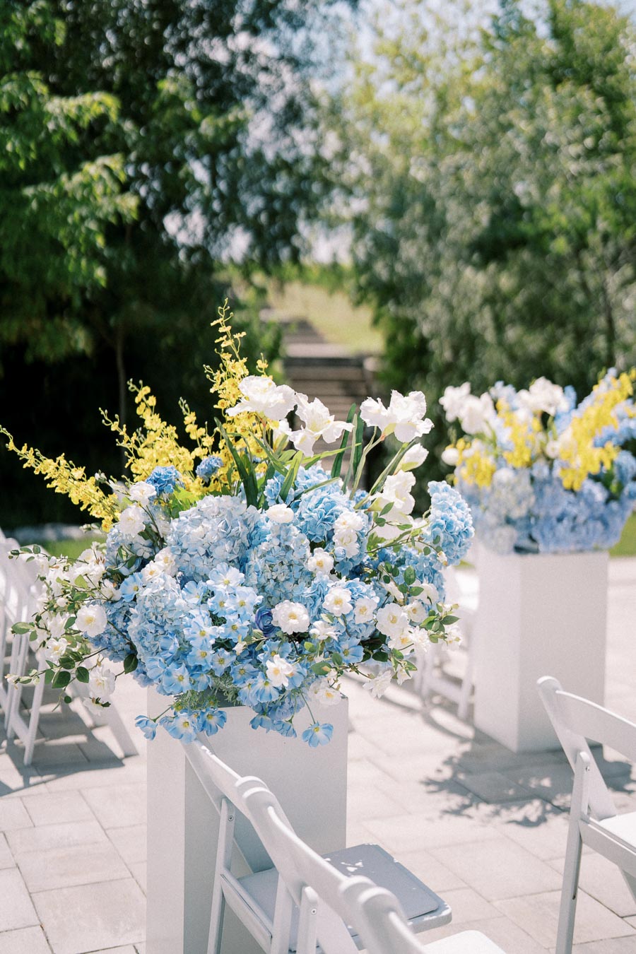 Elegant outdoor wedding ceremony setup featuring vibrant blue and yellow floral arrangements on white pedestals, surrounded by white folding chairs under a lush green backdrop.