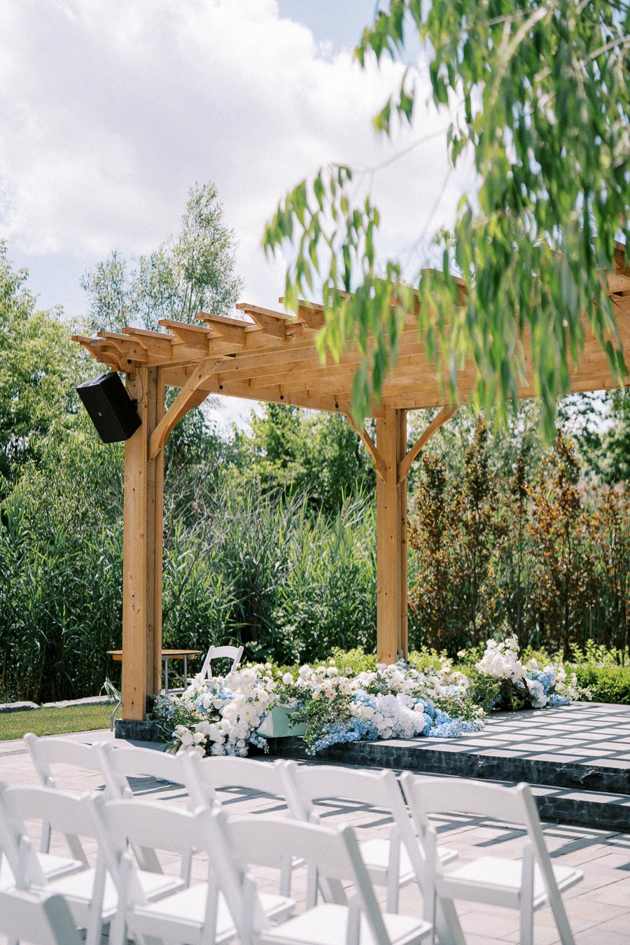 Outdoor wedding ceremony setup, featuring a wooden pergola adorned with white and pastel flowers, surrounded by lush greenery, blue sky, and neatly arranged white chairs.