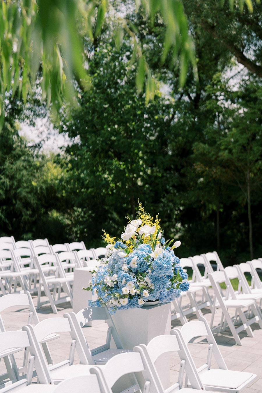 Outdoor wedding ceremony setup with rows of white folding chairs and a large floral arrangement featuring blue and white flowers, surrounded by lush green trees.