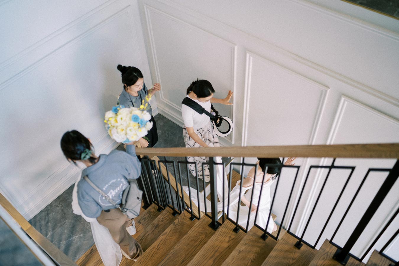Aerial view of a bridal party descending a modern wooden staircase, with an elegant white wall backdrop. One person carries a bouquet of yellow and blue flowers, while the group is dressed in coordinated attire, signifying a wedding preparation moment.