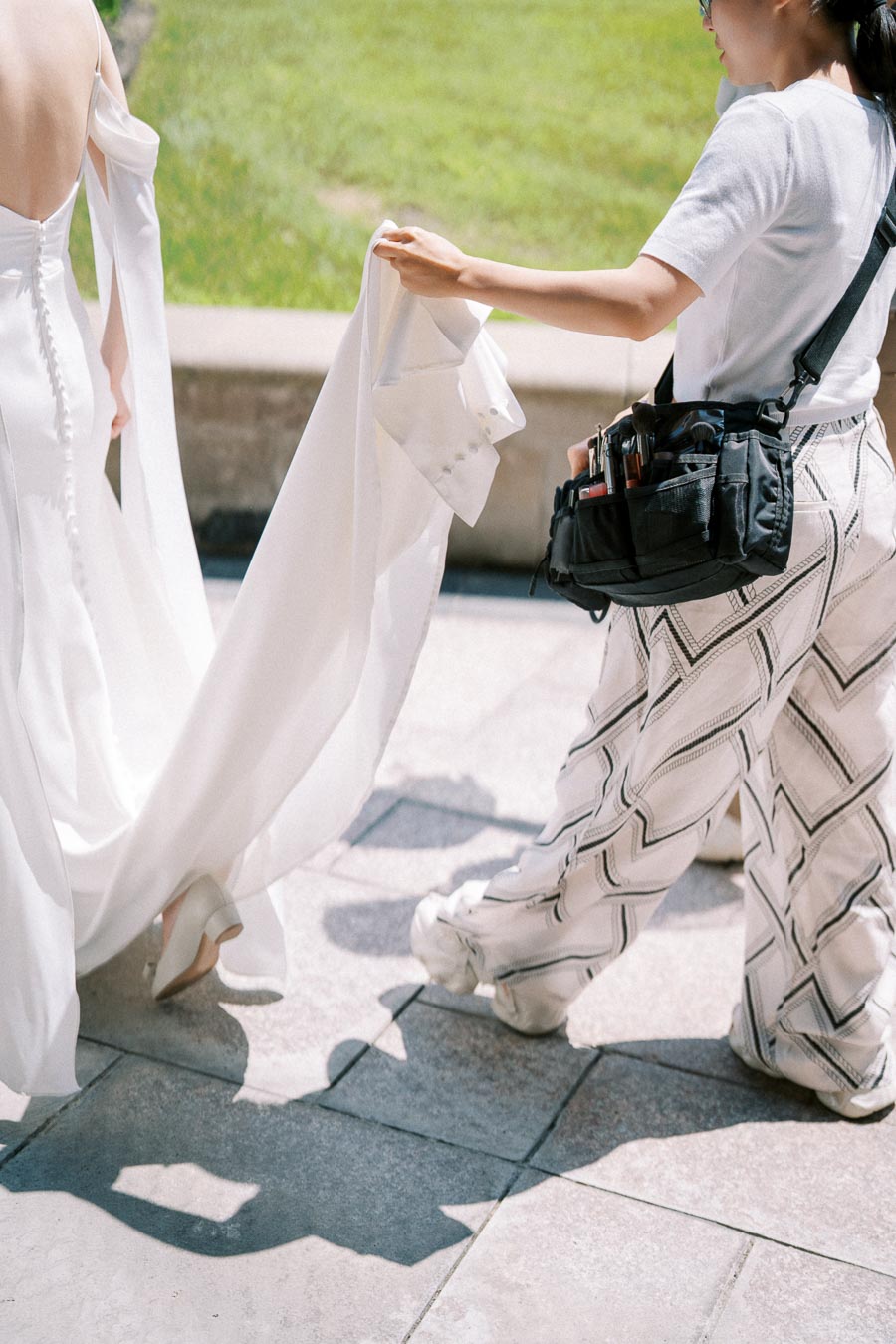 Bride in elegant white wedding dress with long train assisted by a wedding assistant in patterned pants, outdoors on a sunny day.
