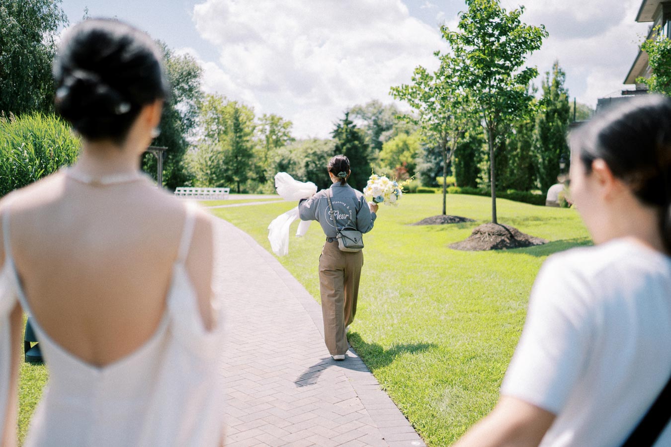 Bride walking in a sunny garden following a person holding flowers, with lush greenery and a pathway leading through a serene outdoor setting.