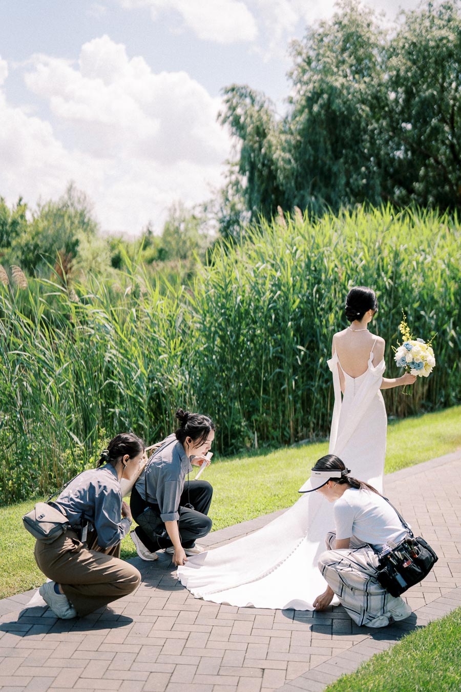 A bride in a white dress stands on a brick path holding a bouquet while three people adjust her train, surrounded by lush greenery and trees under a partly cloudy sky.