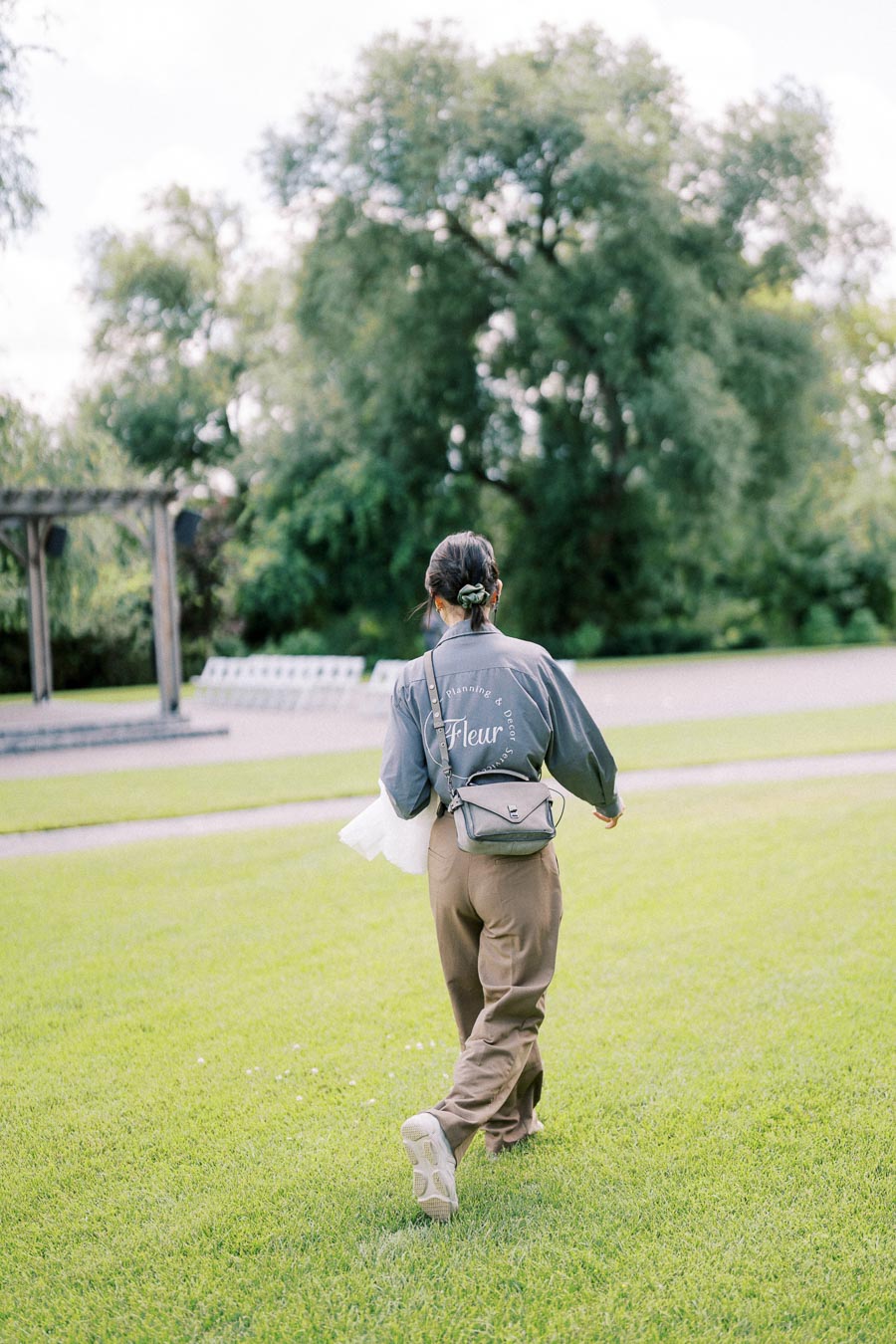 A person walking through a lush green park wearing a gray shirt with text on the back, holding a white cloth and carrying a small crossbody bag.