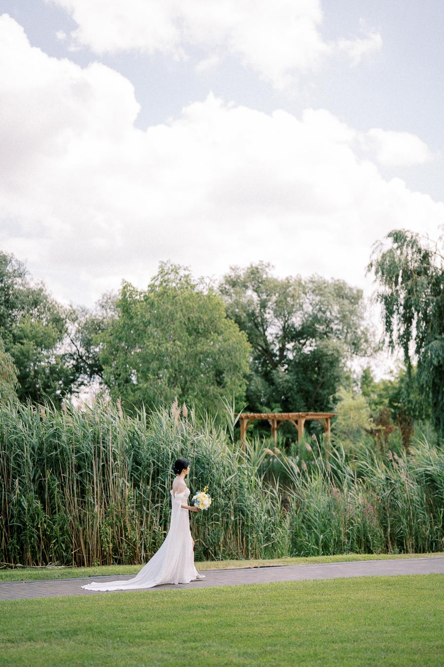 Bride walking through a lush green park holding a bouquet, with tall grass and trees in the background under a partly cloudy sky.