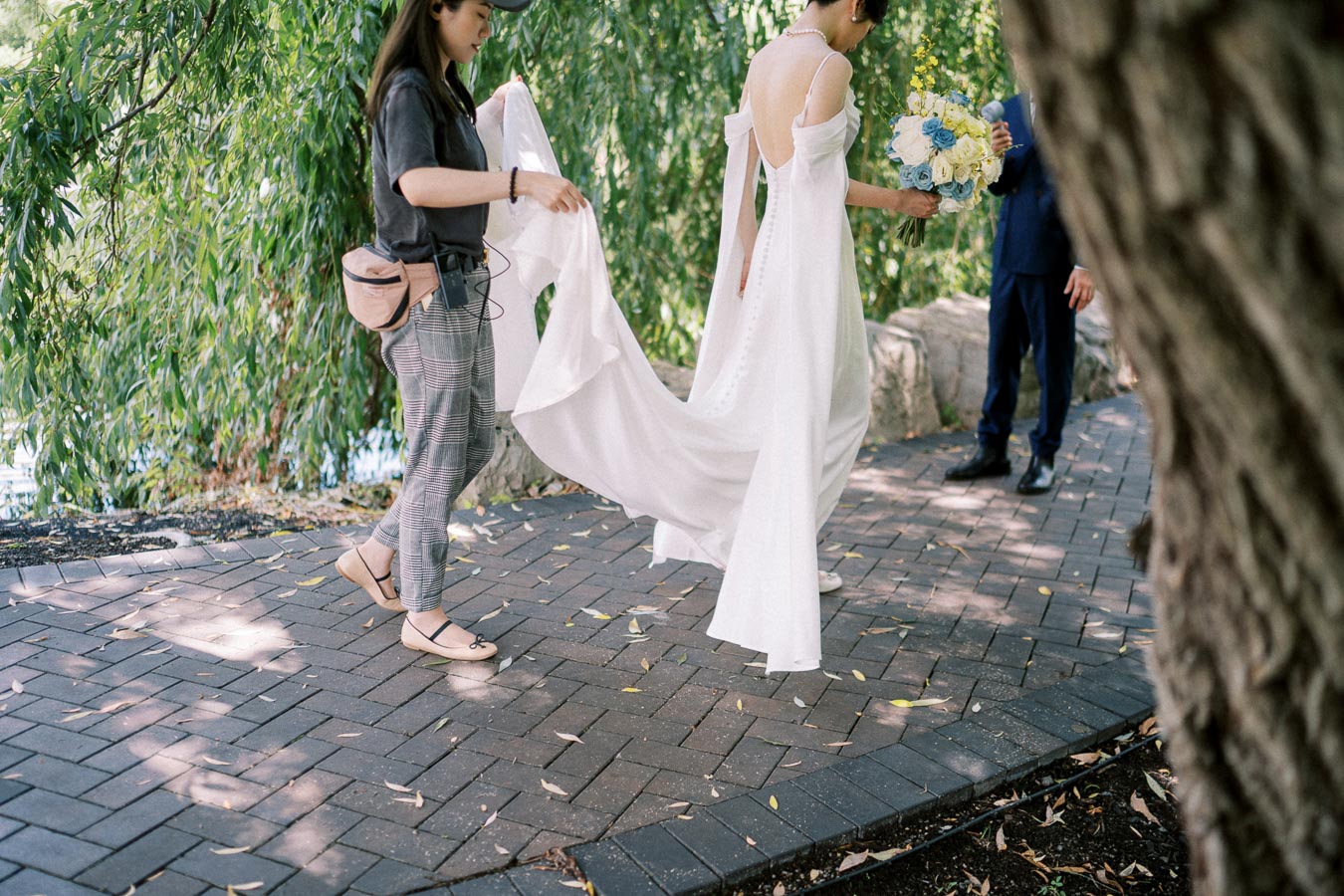 A bride in an elegant white wedding gown and holding a bouquet walks along a brick path, assisted by an attendant, surrounded by lush greenery and accompanied by a man in a suit.