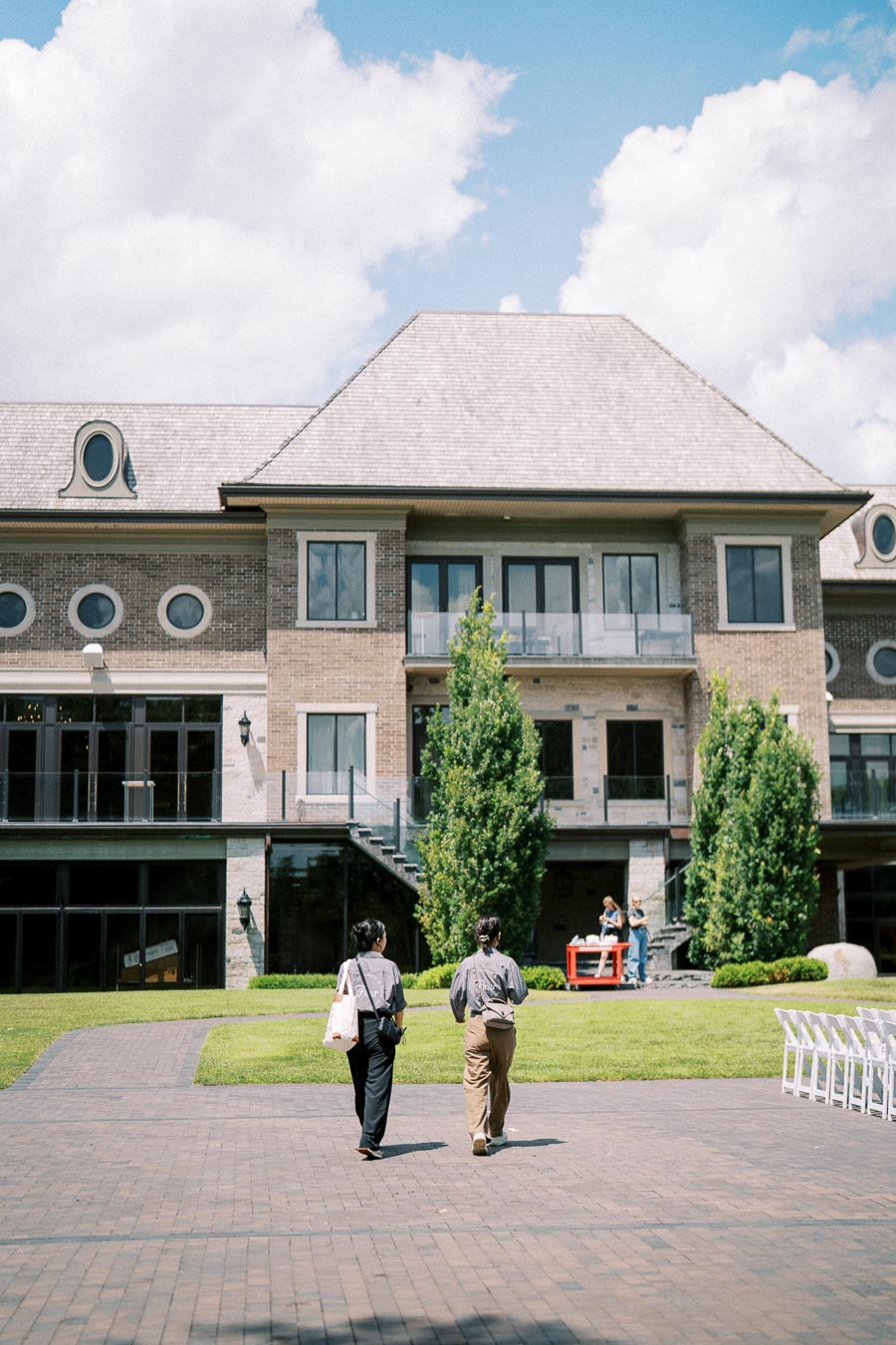 Two people walking towards a large brick building with a pitched roof and multiple windows, set against a clear blue sky with scattered clouds. The foreground features a neatly paved walkway surrounded by green grass and small trees.