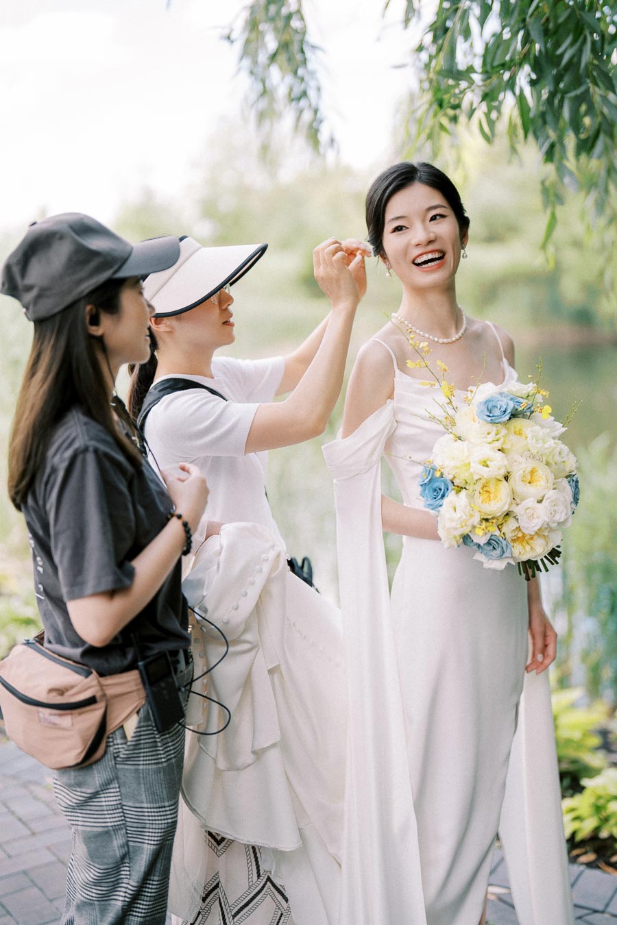 A bride in an elegant white gown holding a bouquet of blue and yellow flowers, surrounded by two women assisting with her attire and accessories in a lush outdoor setting.