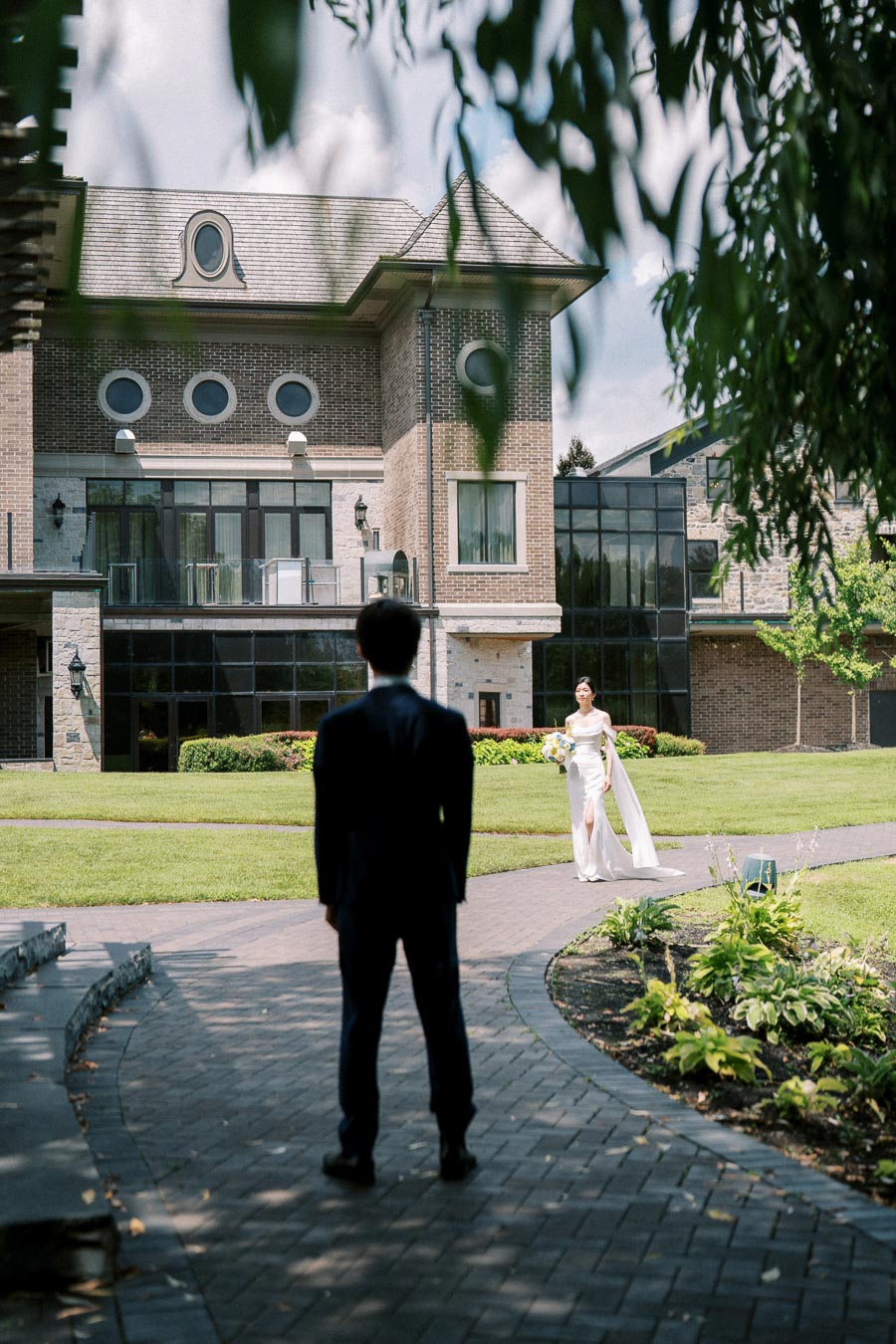 A groom stands in anticipation as a bride in a white dress walks towards him on a brick path in front of a large brick mansion surrounded by greenery on a sunny day.
