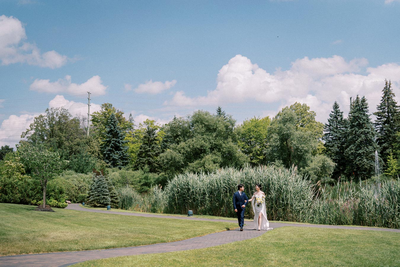 Wedding couple walking on a landscaped path surrounded by lush greenery and trees under a bright blue sky with fluffy white clouds.
