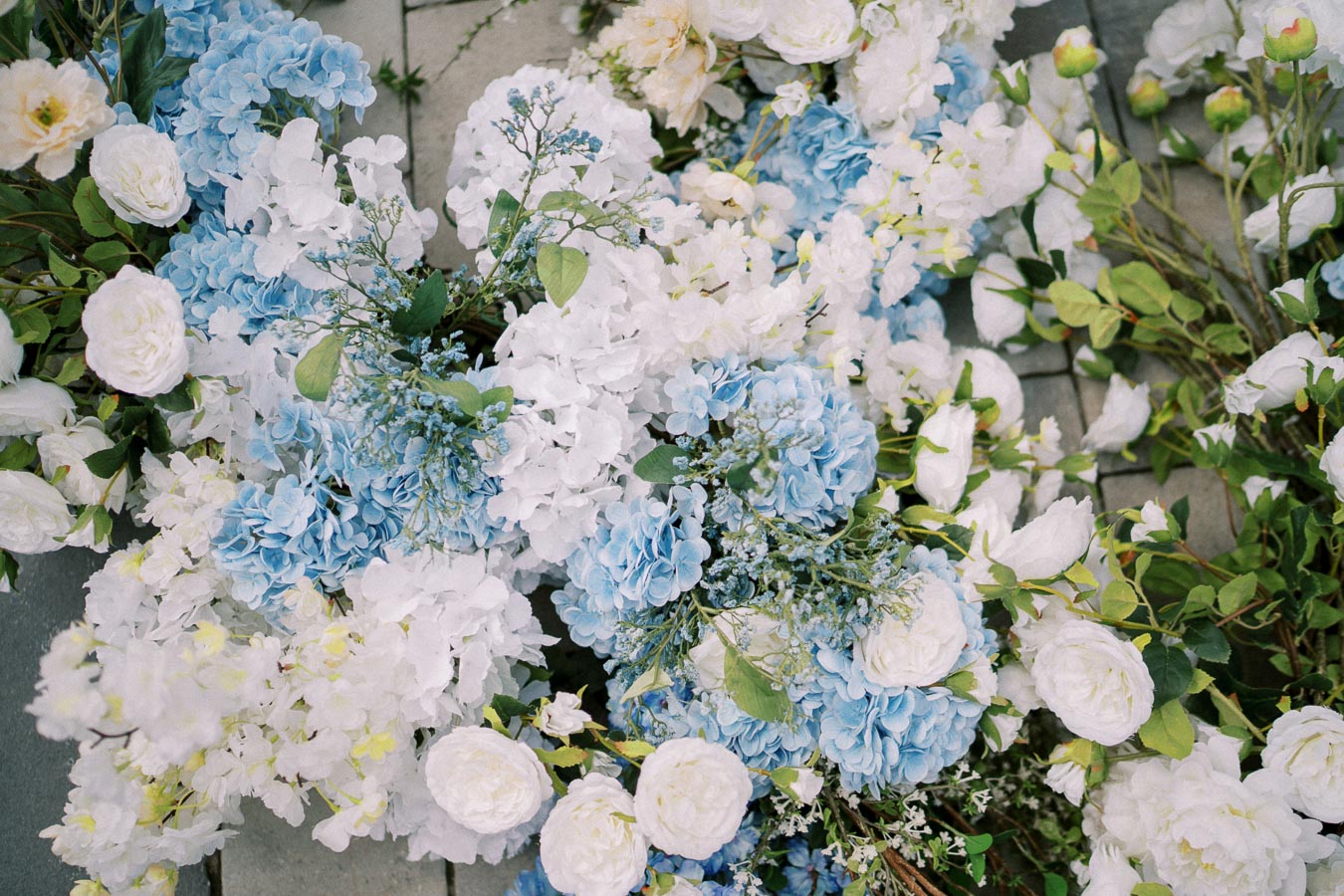 Beautiful arrangement of blue and white hydrangeas and roses with green leaves on a paved surface.