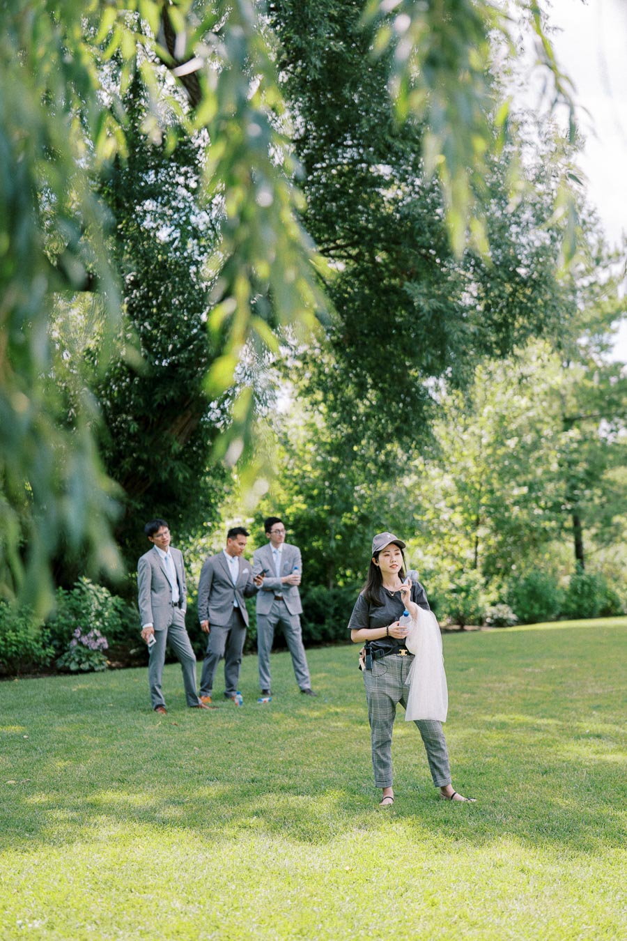 A woman in casual attire stands on a lush green lawn with a group of three men in gray suits in the background under a canopy of trees, creating a serene outdoor setting.