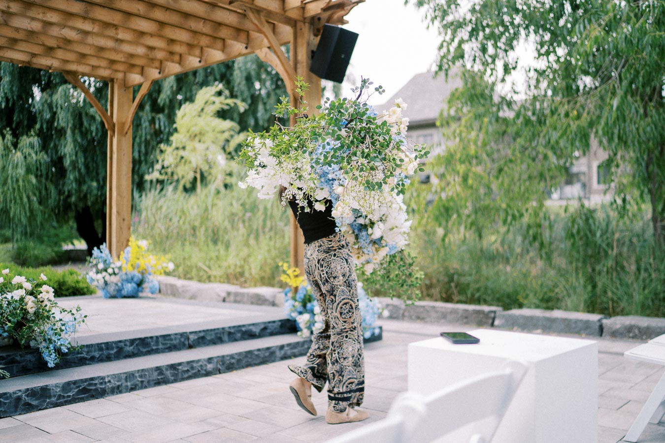Person carrying a large bouquet of blue and white flowers under a wooden pergola in a lush garden setting.
