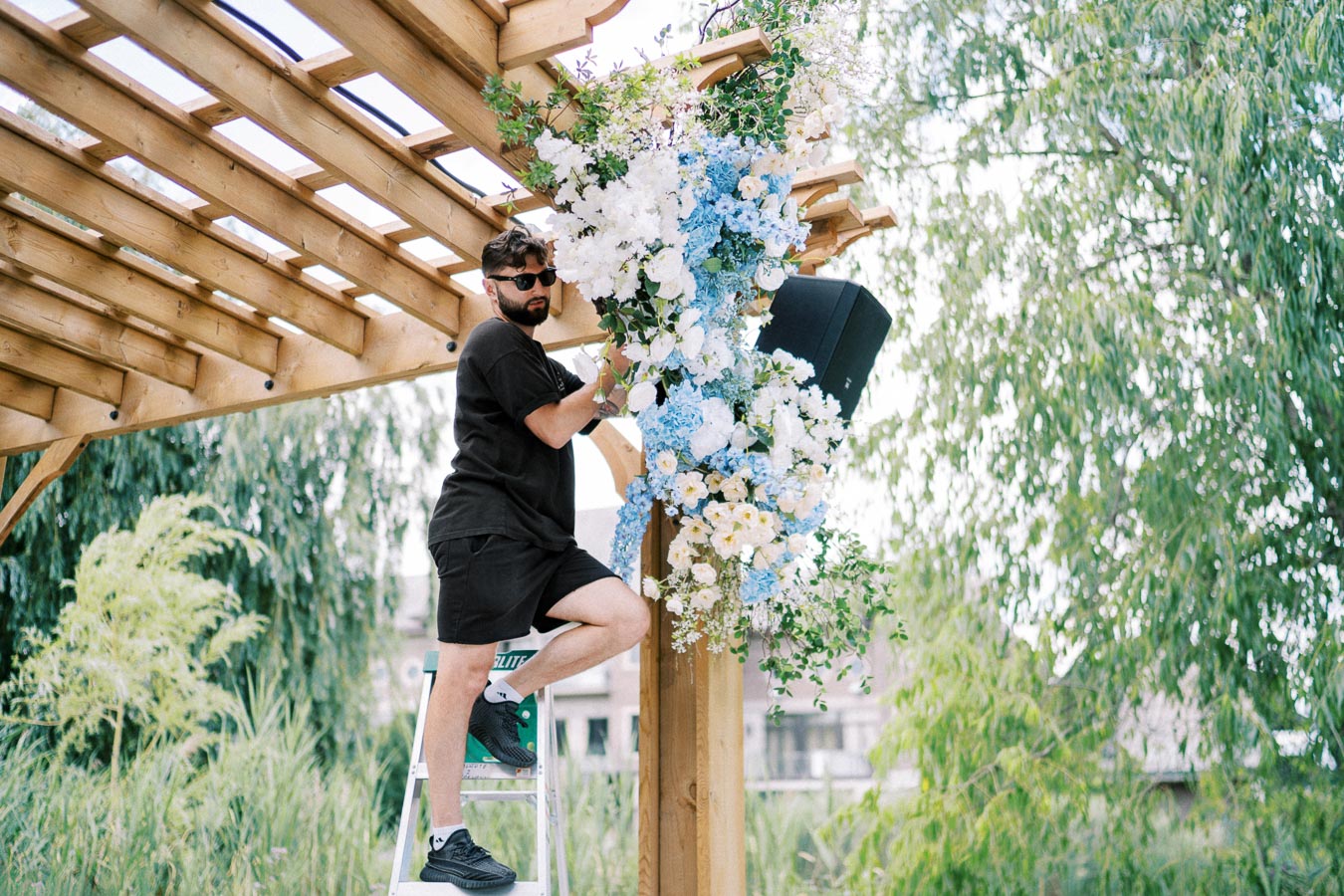 Man in black attire decorating a wooden pergola with blue and white floral arrangements, standing on a ladder, surrounded by lush greenery.