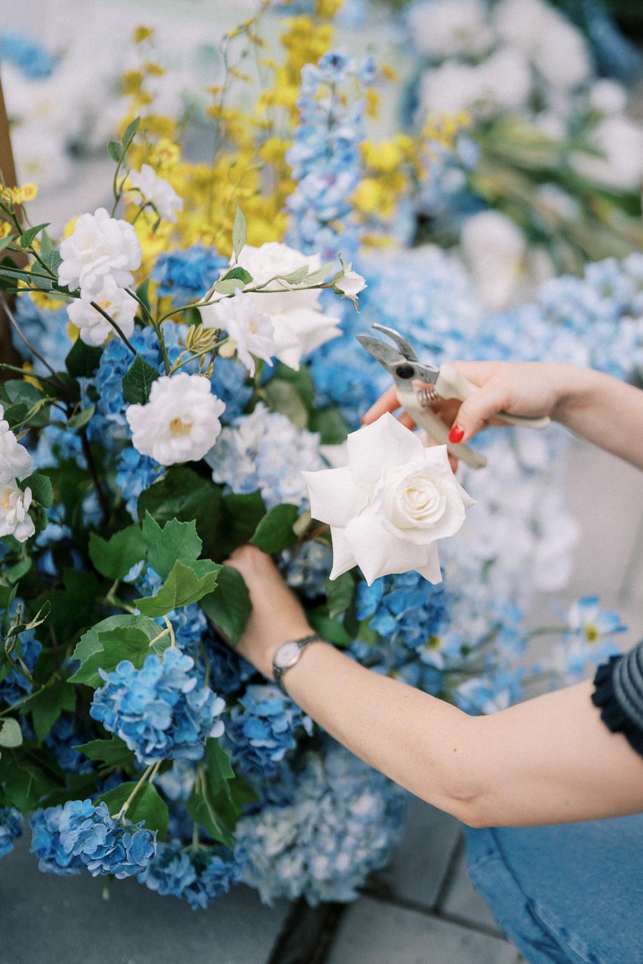 A person arranging a colorful floral bouquet featuring white roses, blue hydrangeas, and yellow accents, using pruning shears.