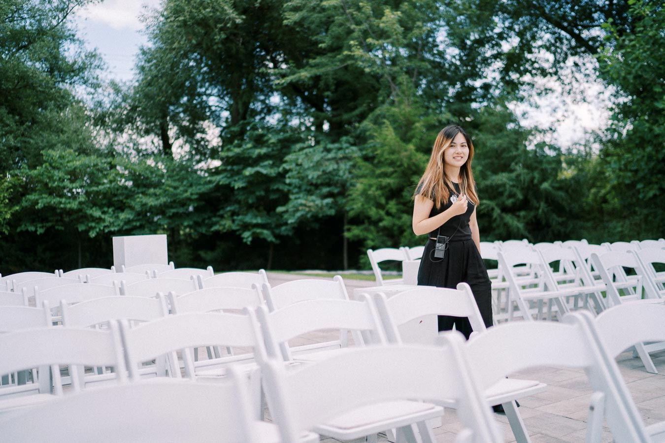 A woman in a black dress stands smiling among rows of empty white chairs set up outdoors, with a lush green forest in the background, suggesting an event or ceremony setting.
