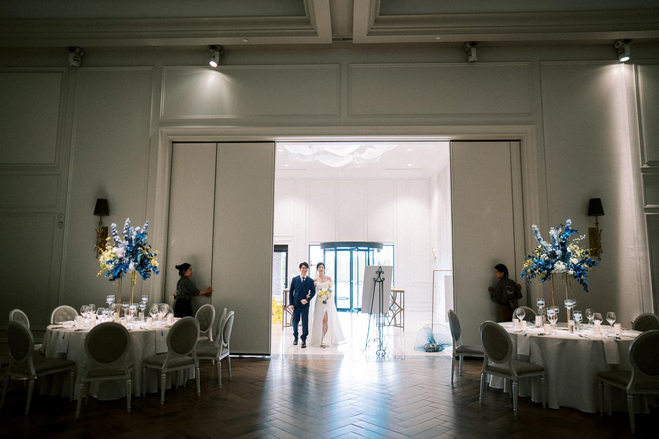 Elegant wedding entrance with a couple walking into a beautifully decorated reception hall, featuring round tables with white tablecloths, ornate floral arrangements in blue and yellow, and stylish lighting, creating a sophisticated ambiance.