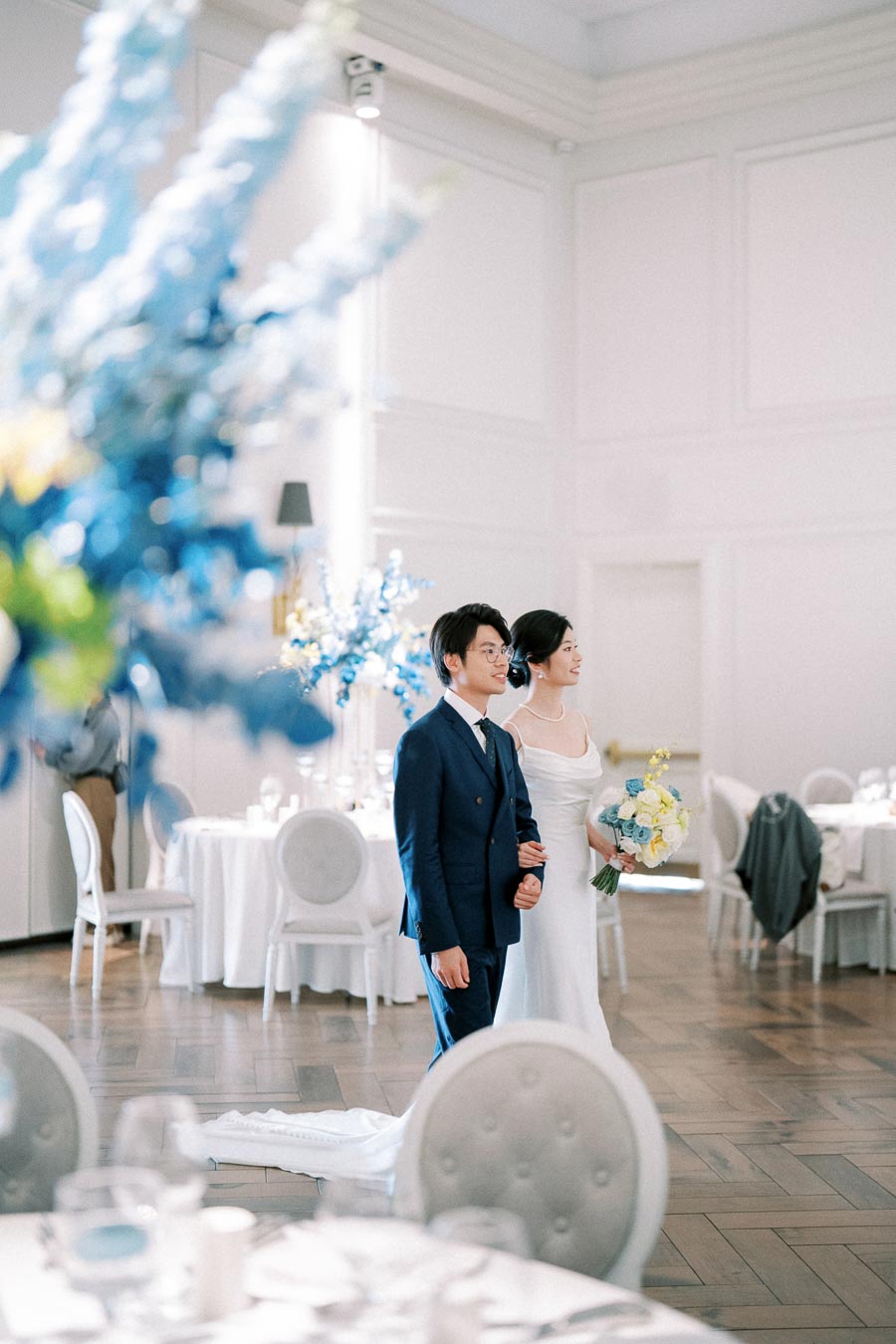 A newlywed couple walks arm in arm in an elegantly decorated wedding reception hall, with blue and white floral arrangements in the foreground and round tables set for guests. The bride holds a bouquet of yellow and blue flowers, wearing a white dress, while the groom is in a dark blue suit.