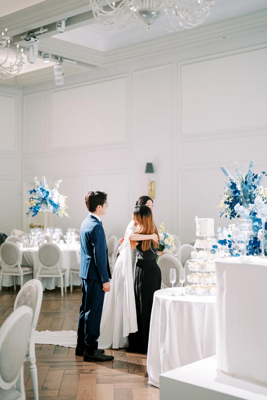 Elegant wedding reception with bride and groom interacting with guest. The room features sophisticated white decor and blue floral arrangements, surrounded by tastefully set dining tables.