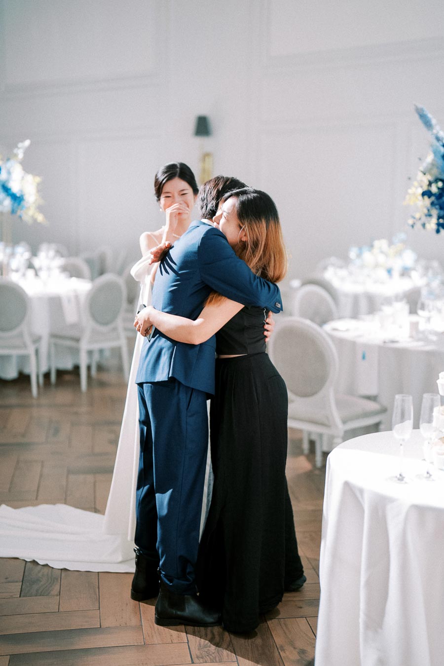 Wedding celebration with guests embracing, featuring a joyful scene in a bright, elegant reception setting. Tables are adorned with white tablecloths and floral centerpieces, creating a warm, festive atmosphere.