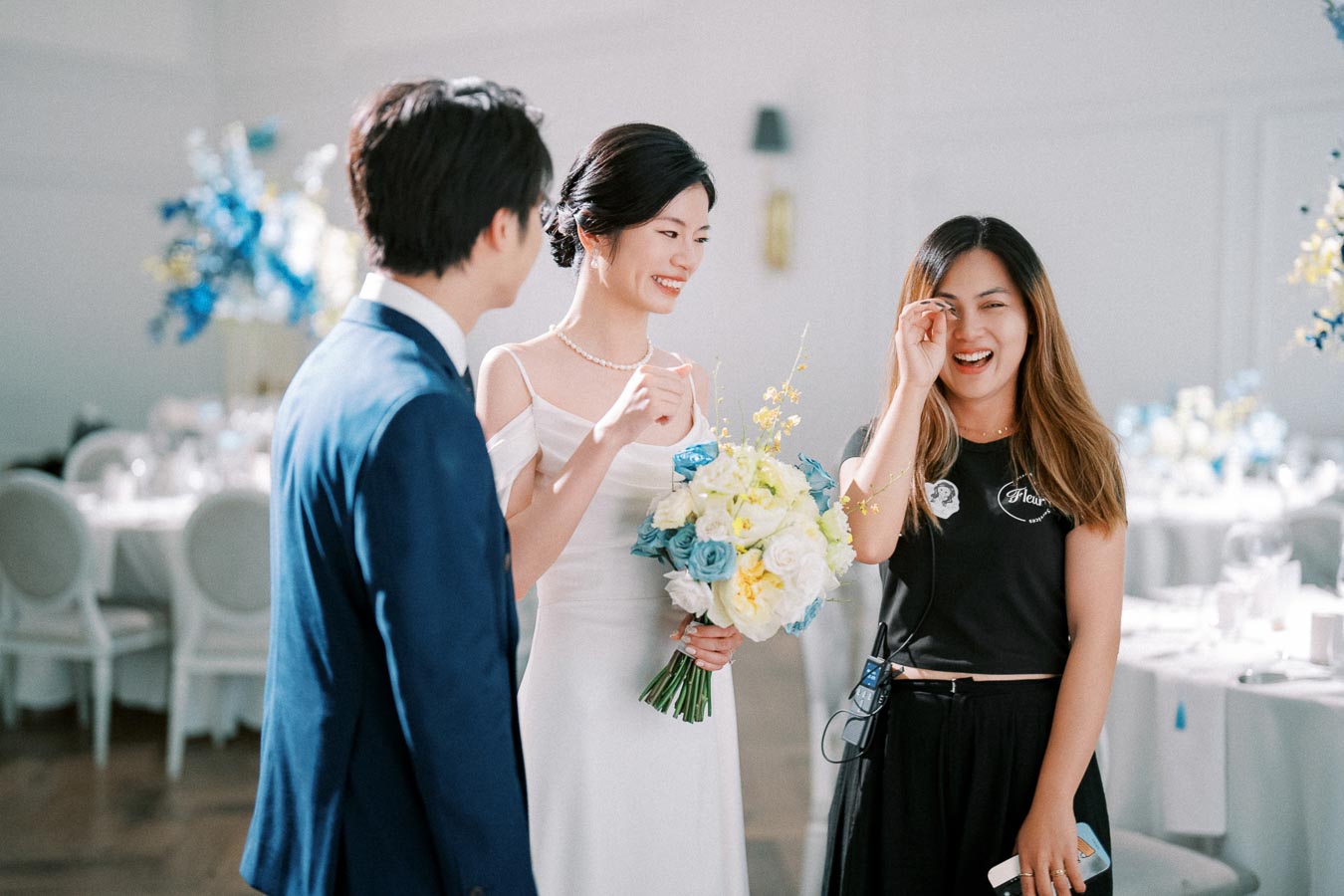A bride in a white wedding dress holding a bouquet smiles at a man in a blue suit and a woman in a black shirt, inside a decorated wedding venue.
