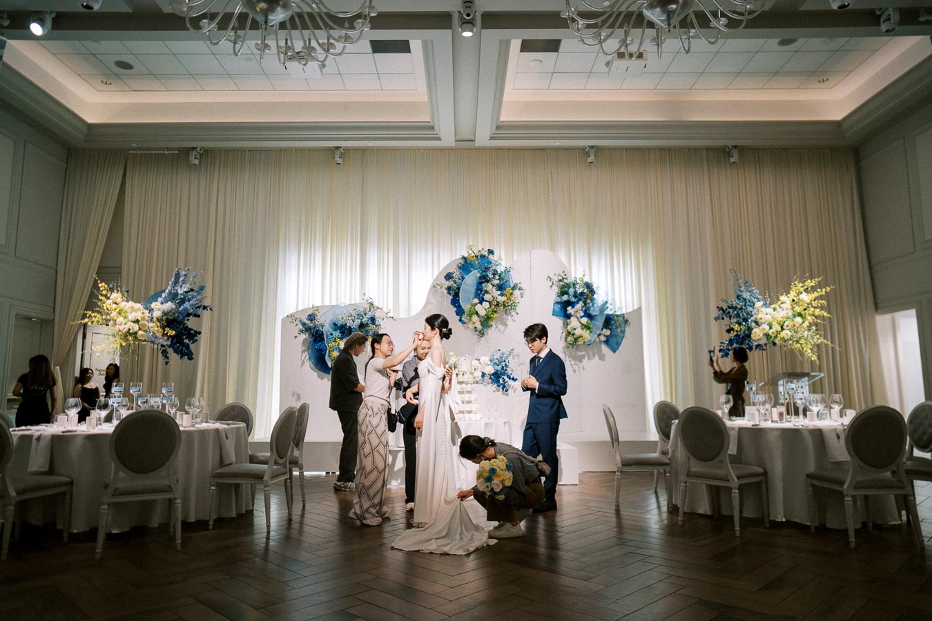 Elegant wedding setup with bride and groom getting ready on stage, surrounded by floral arrangements in a decorated reception hall.
