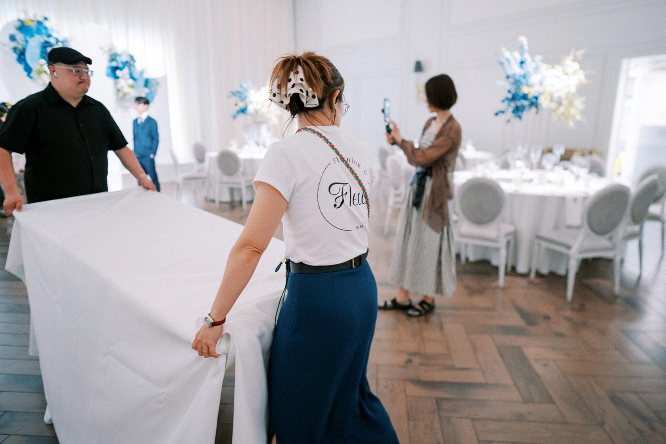 Event planners preparing a venue, arranging a table with a white tablecloth in a beautifully decorated room with floral arrangements and elegantly set tables.