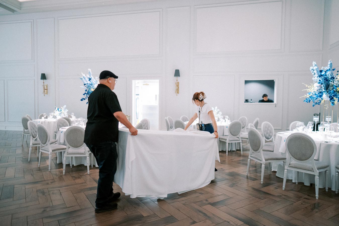 Elegant event hall setup with two individuals arranging a white tablecloth on a round dining table, surrounded by elegantly set tables with chairs and blue floral arrangements, showcasing event preparation and decor.