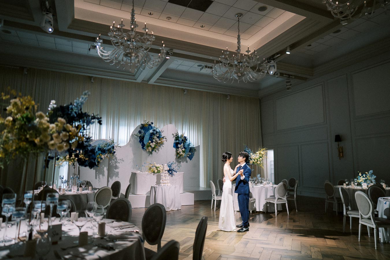 A bride and groom share their first dance in an elegantly decorated ballroom, featuring chandeliers, floral arrangements in blue and white hues, and set tables for a wedding reception.