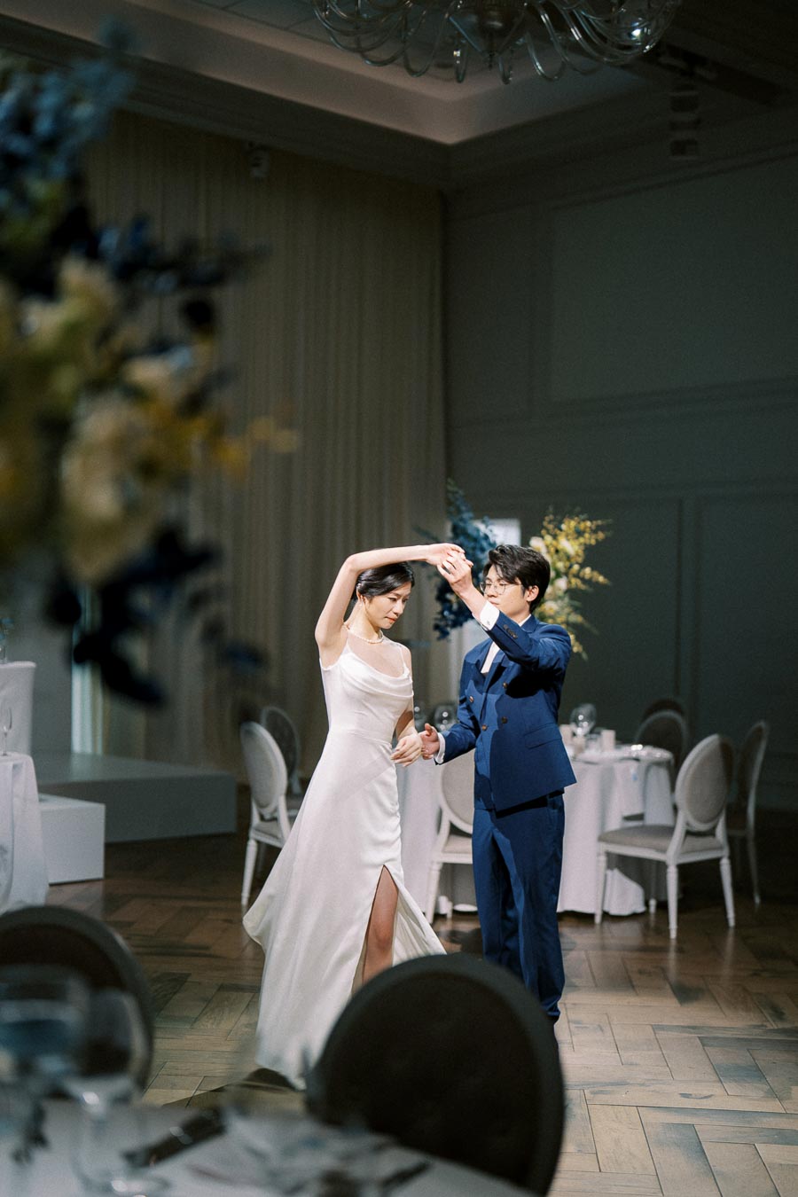 Bride and groom share a romantic first dance at an elegant indoor wedding reception, showcasing a beautifully decorated venue with round tables and floral arrangements.