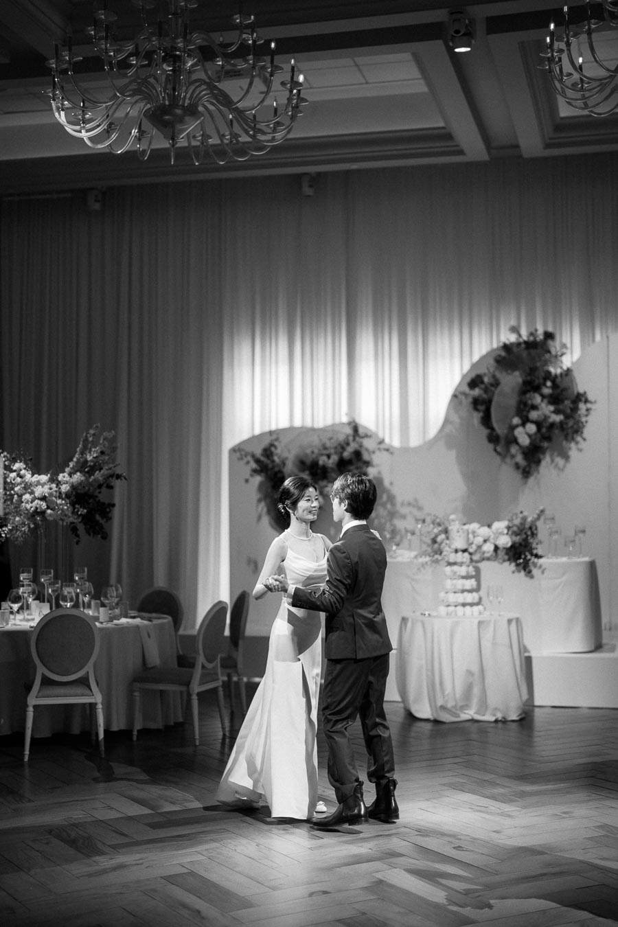 Black and white image of a couple sharing their first dance at an elegant wedding reception, with a beautifully decorated cake table and floral arrangements in the background.