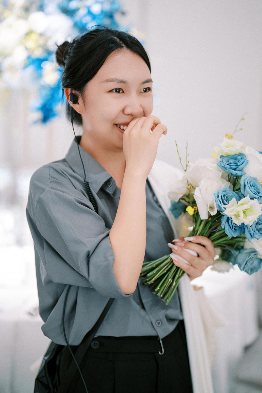 Young woman smiling and holding a bouquet of blue and white flowers, wearing a blue shirt, with a bright, blurred floral background.
