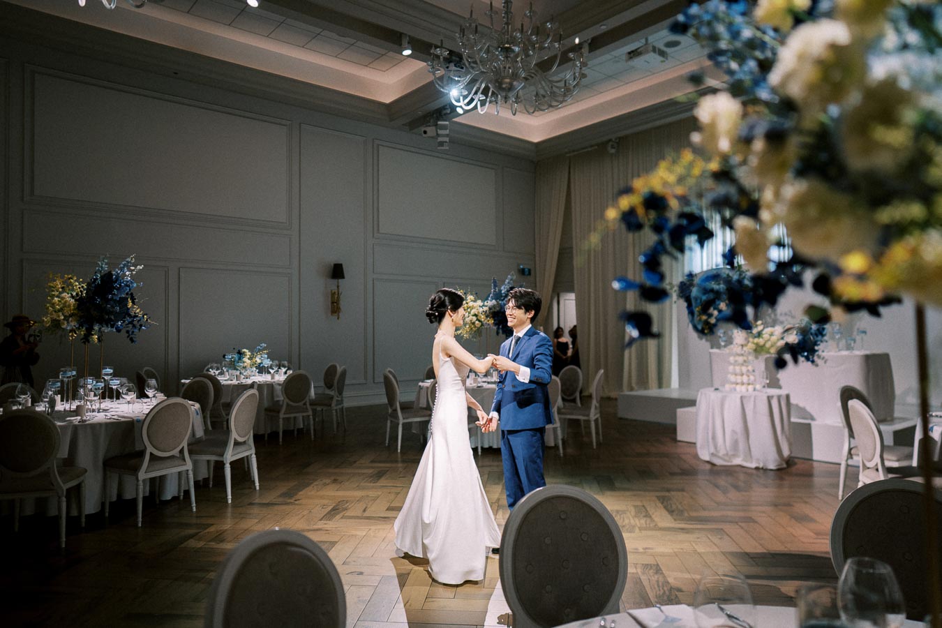 A couple sharing a romantic dance in an elegant wedding reception hall decorated with blue and yellow floral arrangements.