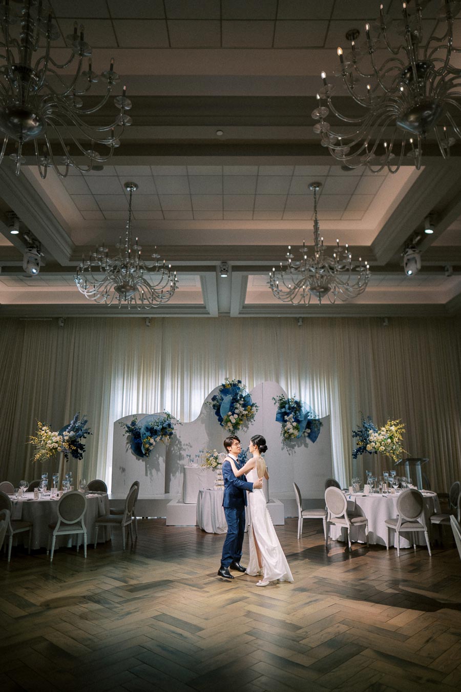 Elegant couple dancing in a beautifully decorated wedding ballroom with chandeliers and floral arrangements.