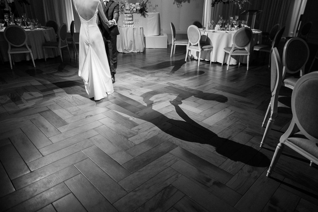 Black and white photo of a couple dancing closely on a wooden floor at a wedding reception, casting a dramatic shadow.