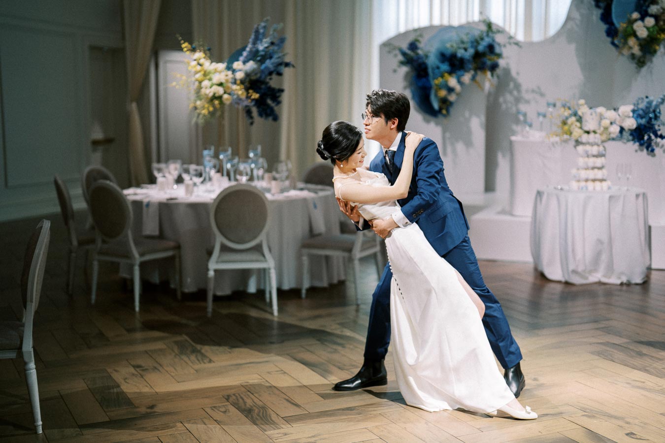 A couple dancing at a wedding reception, with the groom in a blue suit and the bride in a white gown. The elegantly decorated venue features floral arrangements in blue and white, with a round table set for guests in the background.