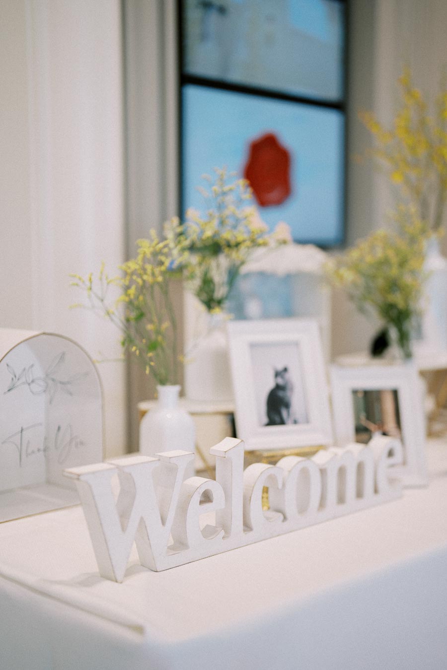 Alt text: Elegant welcome table with a white 'Welcome' sign, framed photos, potted flowers, and decorative vases, set against a soft, neutral backdrop perfect for a wedding or event entrance display.