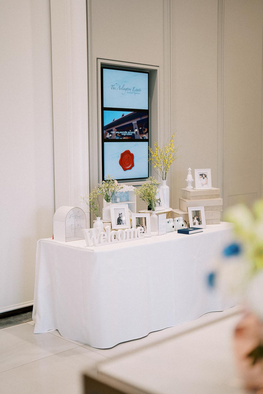 Elegant welcome table setup with decorative frames and flowers at a special event, featuring a white tablecloth and a warm, inviting atmosphere.