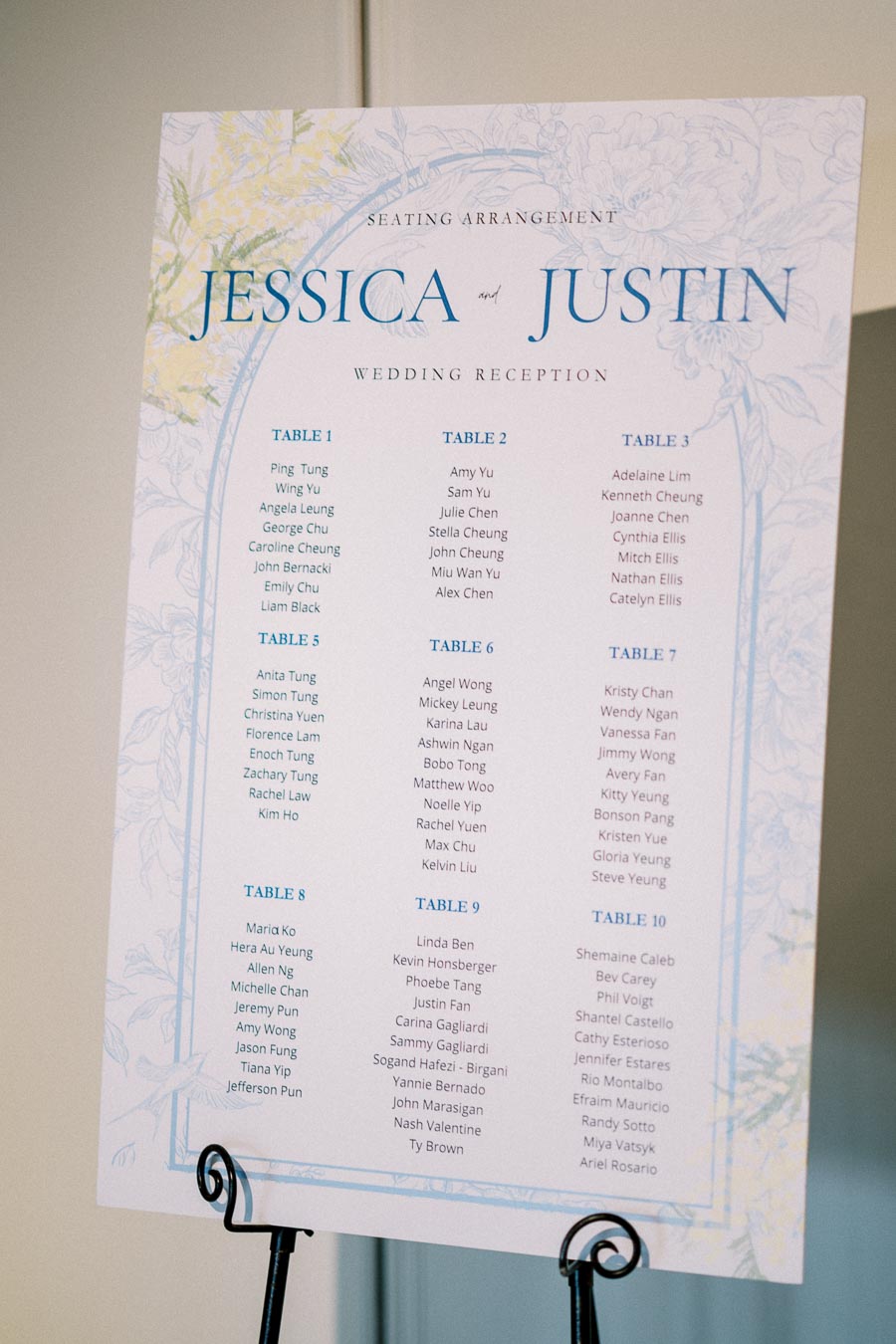 Elegant wedding reception seating chart for Jessica and Justin, featuring guest names organized by tables, adorned with floral designs, displayed on a stand.