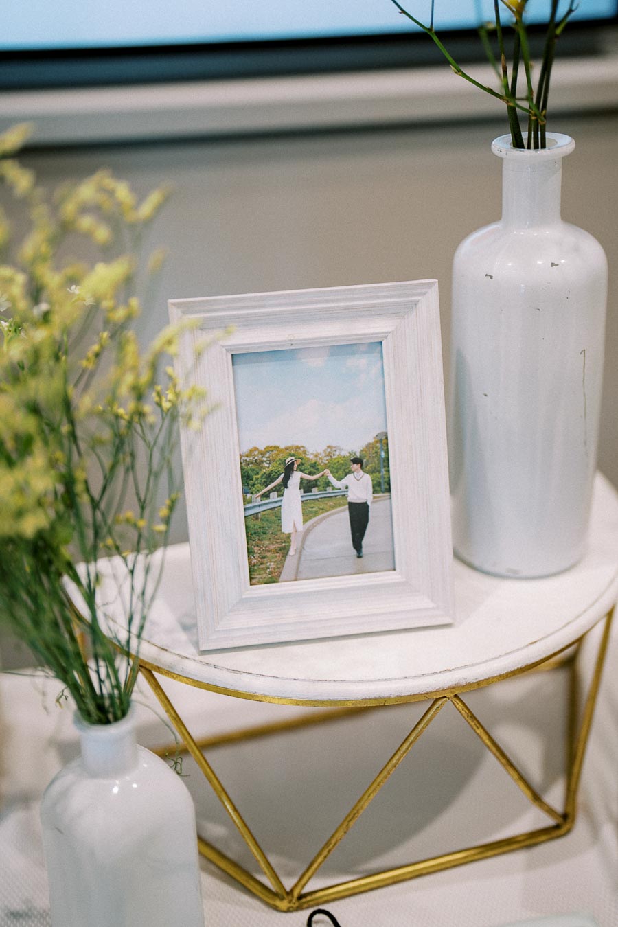 Framed photo of a couple holding hands outdoors, displayed on a white table with decorative elements such as yellow flowers and white ceramic vases.