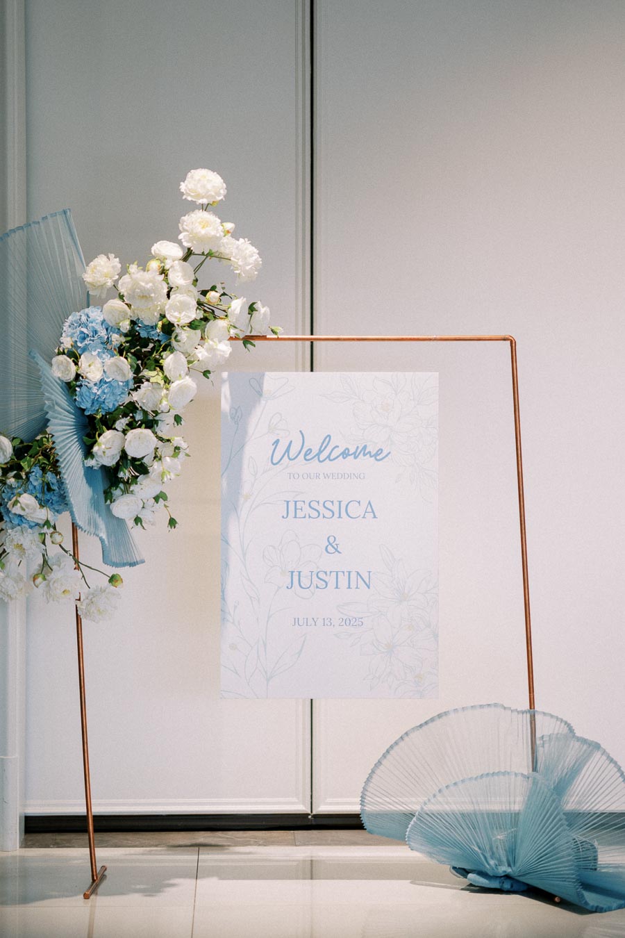 Elegant wedding welcome sign with floral arrangement, featuring white and blue flowers and decorative accents, announces wedding of Jessica and Justin on July 13, 2025.