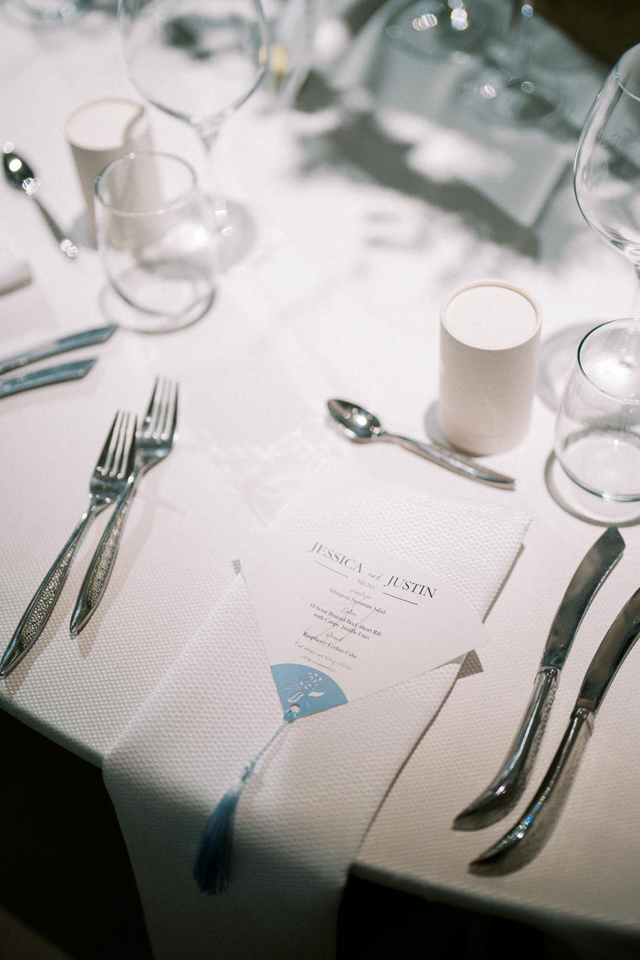 Elegant wedding table setting with a white tablecloth, featuring a menu card placed in a neatly folded napkin, surrounded by silverware, wine glasses, and a soft-lit ambiance.