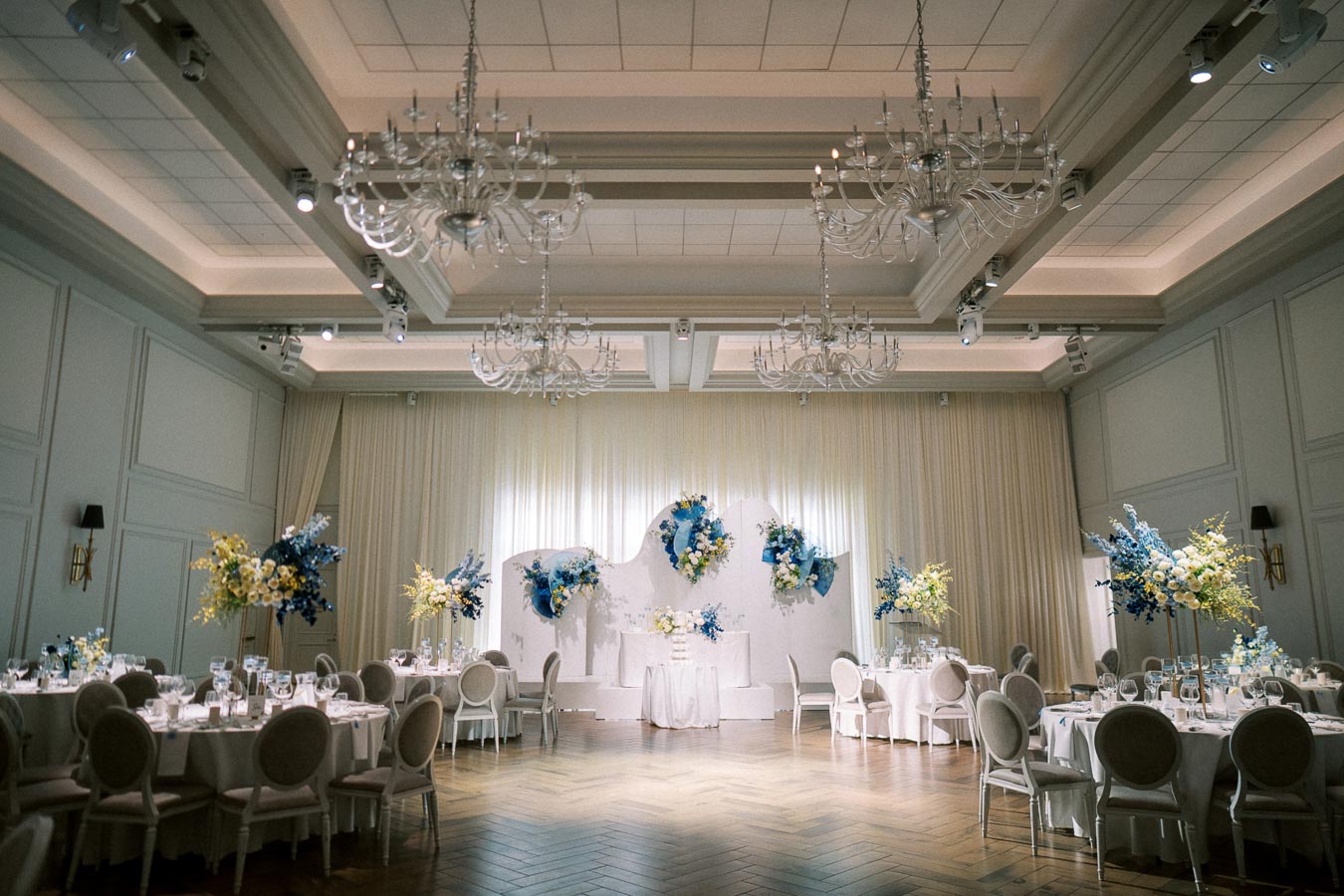 Elegant wedding reception hall with chandeliers and round tables adorned with white tablecloths and decorative yellow and blue floral arrangements.